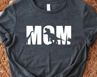 Puede incluir: Una camiseta gris oscuro con la palabra "MOM" impresa en blanco. Las letras están delineadas con siluetas de dinosaurios.