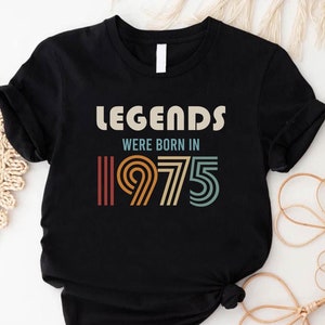 May include: Black t-shirt with the text "Legends were born in 1975" in a retro style font. The numbers are in a rainbow color scheme.