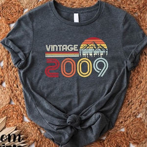 May include: A dark gray t-shirt with a vintage-style graphic design featuring a sunset over mountains and the text "Vintage 2009".