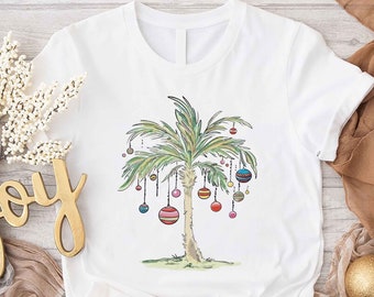 May include: White t-shirt featuring a watercolor illustration of a palm tree decorated with colorful ornaments. The design evokes a tropical Christmas theme. The shirt is made of a soft, comfortable material, perfect for casual wear.