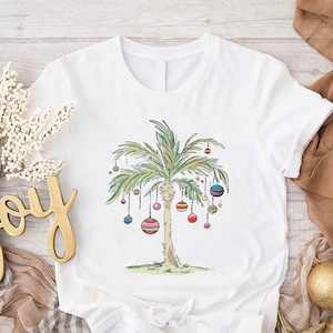 May include: White t-shirt featuring a watercolor illustration of a palm tree decorated with colorful ornaments. The design evokes a tropical Christmas theme. The shirt is made of a soft, comfortable material, perfect for casual wear.