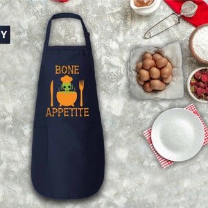 May include: Navy blue apron with a white pocket and orange text that reads "BONE APPETITE". A cartoon skeleton chef is cooking in a cauldron.