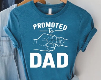 Puede incluir: Una camiseta azul turquesa con texto blanco que dice "Promovido a papá" con un gráfico de puñetazos.