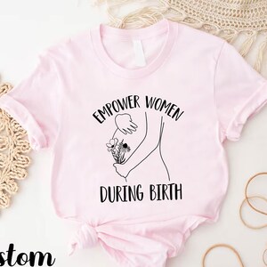 May include: Pink t-shirt with a black graphic design that says "Empower Women During Birth" and a line drawing of a pregnant person holding flowers.