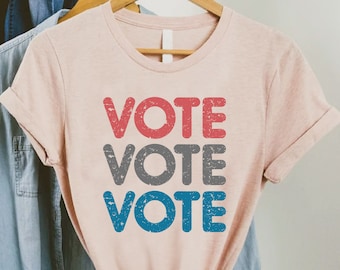 May include: A pink t-shirt with the word "VOTE" repeated three times in red, gray, and blue. The text has a distressed, vintage look.