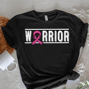 May include: Black t-shirt with the word "WARRIOR" in white block letters. A pink ribbon with two fists is in the center of the word. The shirt is on a white surface with a woven basket and a white mug.