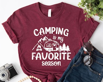 May include: A maroon t-shirt with the text "Camping is my favorite season" in white. The design includes a camper, mountains, trees, and a campfire.