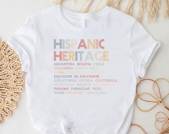 Op de afbeelding: Wit t-shirt met de tekst "HISPANIC HERITAGE" in pastelkleuren en een lijst met landen eronder. Het shirt heeft ook de tekst "Custom DTFPrints" onderaan.