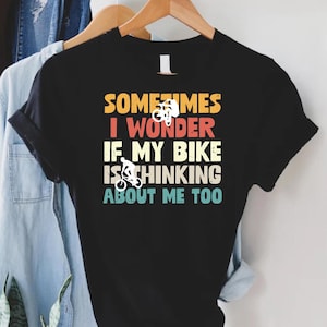 May include: Black t-shirt with the text "SOMETIMES I WONDER IF MY BIKE IS THINKING ABOUT ME TOO" in a retro font. The design includes white bicycle illustrations. The shirt is hanging on a wooden hanger with other clothing.