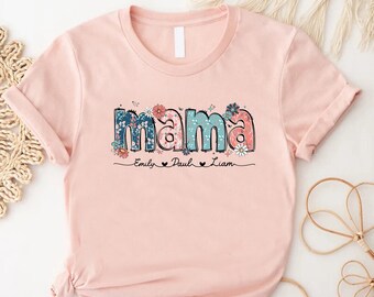 May include: A light pink t-shirt with the word "mama" in a floral design. The letters are in blue, pink, and white with floral accents. Below the word "mama" are the names "Emily", "Paul", and "Liam".