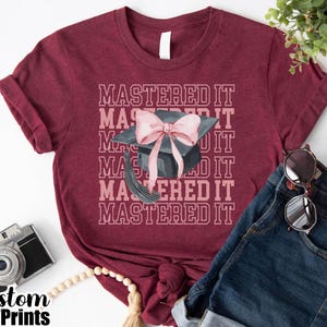 May include: Burgundy t-shirt with the text "MASTERED IT" repeated vertically. A graduation cap with a pink bow is centered on the shirt. Sunglasses and denim shorts are next to the shirt. A vintage camera and a string of beads are also in the image.