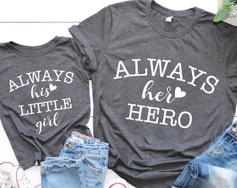 Puede incluir: Dos camisetas grises con texto blanco. La camiseta más grande dice "ALWAYS her HERO" con un pequeño símbolo de corazón. La camiseta más pequeña dice "ALWAYS his LITTLE girl" con un pequeño símbolo de corazón.