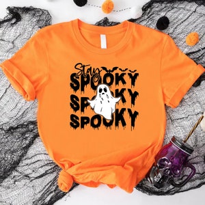 May include: Orange t-shirt with the words "Stay Spooky" in black, along with a white ghost illustration. The word "Spooky" is repeated three times. Bats are above the first word. A Halloween-themed shirt.