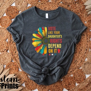 May include: A dark gray t-shirt with a colorful flower graphic and the text "Vote like your daughter's rights depend on it".