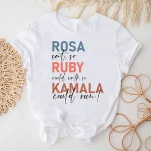 May include: White t-shirt with a graphic design featuring the text "ROSA sat so RUBY could walk, so KAMALA could run!" in various colors.