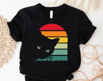 May include: Black t-shirt with a retro sunset graphic and a black cat peeking out from behind it. The sunset is in shades of red, orange, yellow, and green.