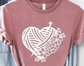 May include: A pink t-shirt with a white graphic of a heart-shaped ball of yarn with a needle and flowers.