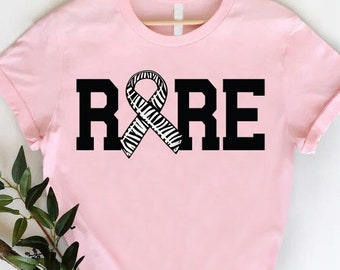 Puede incluir: Camiseta rosa con una cinta negra y la palabra "RARE" impresa en ella.