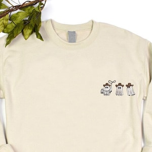 a white sweater with embroidered three cowboy boo ghosts on it