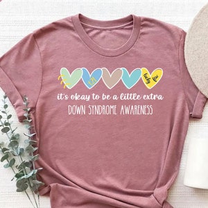 May include: Heather mauve t-shirt with a row of five colorful hearts, each with a different design. The text "it's okay to be a little extra DOWN SYNDROME AWARENESS" is printed below the hearts.