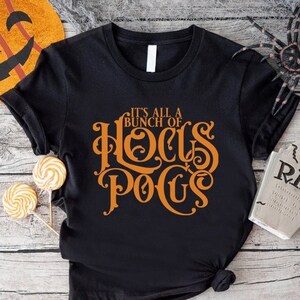 May include: Black t-shirt with the orange text "It's All a Bunch of Hocus Pocus". The shirt is styled with rolled-up sleeves and a knotted hem. Halloween-themed decorations include a pumpkin, spider, and tombstone.