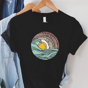 May include: Black t-shirt with a graphic of a stylized sun setting over a wave. The sun is yellow, the wave is blue and green, and the background is a peach and pink gradient.