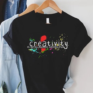 May include: Black t-shirt with the words "creativity is intelligence having fun" in white. The shirt has colorful paint splatter designs. Other clothing items, including a denim shirt and a brown hat, are visible in the background.