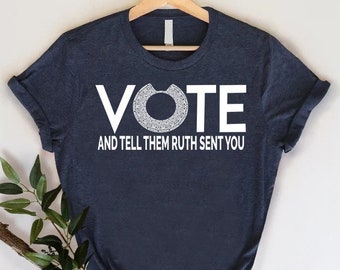 Puede incluir: Una camiseta azul marino con texto blanco que dice "VOTE" con un diseño circular dentro de la "O". Debajo de la palabra "VOTE" está el texto "AND TELL THEM RUTH SENT YOU".