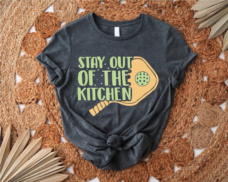 May include: A dark grey t-shirt with the text "Stay Out of the Kitchen" printed on it in a light green and yellow font. A yellow pickleball paddle is behind the text.