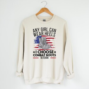 May include: A beige sweatshirt with the text "Any girl can wear heels * I choose combat boots * Veteran" and an American flag with a pair of combat boots on it.