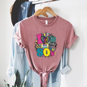 May include: A pink t-shirt featuring a graphic design that reads "I love the 80's" with a heart, a cassette tape, a Rubik's Cube, a disco ball, and other 80s-themed icons.