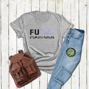May include: A gray t-shirt with the text "FU&infin; STOMACH CANCER" printed on it. The shirt is laid out on a white wooden surface with a pair of blue jeans and a brown leather backpack.