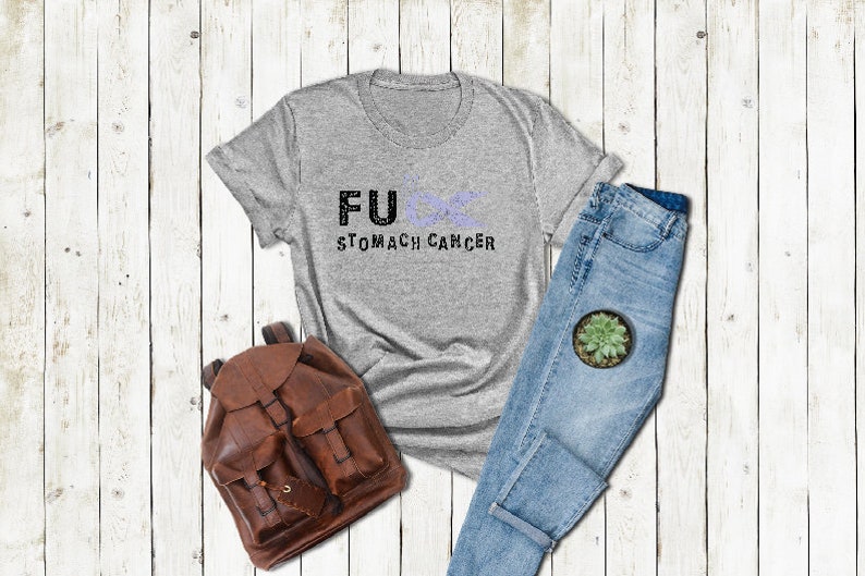 May include: A gray t-shirt with the text "FU&infin; STOMACH CANCER" printed on it. The shirt is laid out on a white wooden surface with a pair of blue jeans and a brown leather backpack.