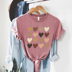 May include: A pink t-shirt with a design of 16 hearts in different shades of brown, representing skin tones.