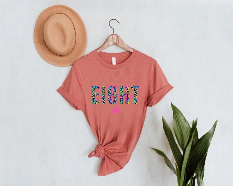 May include: A rust-coloured t-shirt with the word "EIGHT" printed in black, outlined in rainbow colours, and a pink heart below. The letters are filled with a leopard print pattern.