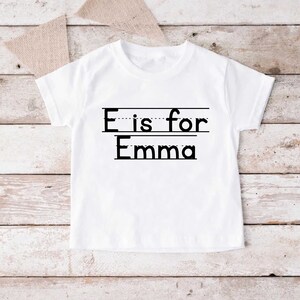 May include: White t-shirt with black text that says "E is for Emma" with a dotted line under the "E" and a solid line under "Emma".