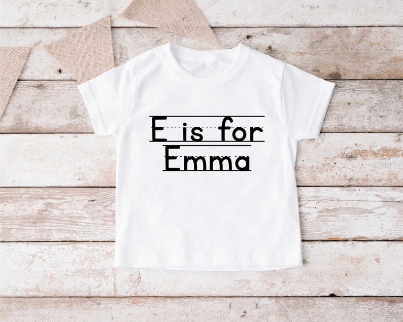May include: White t-shirt with black text that says "E is for Emma" with a dotted line under the "E" and a solid line under "Emma".