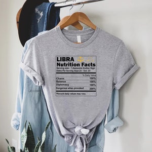May include: Gray t-shirt with a graphic of a nutrition facts label for the zodiac sign Libra. The label lists the following: Charm, Balance, Diplomacy, and Dangerous when provoked. The label states that the daily value for each is 100% except for Dangerous when provoked, which is 200%.