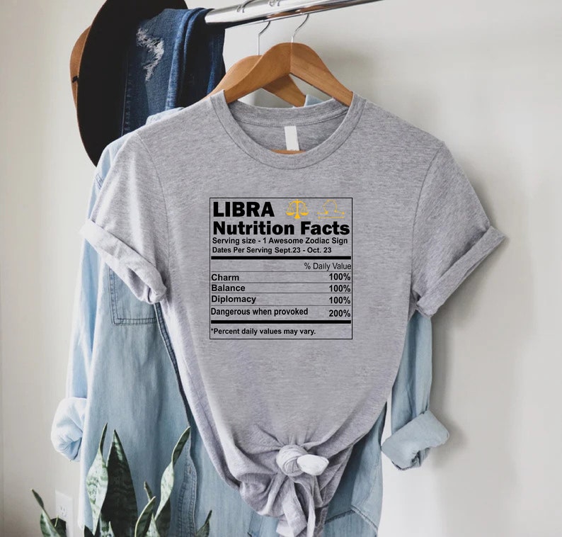 May include: Gray t-shirt with a graphic of a nutrition facts label for the zodiac sign Libra. The label lists the following: Charm, Balance, Diplomacy, and Dangerous when provoked. The label states that the daily value for each is 100% except for Dangerous when provoked, which is 200%.