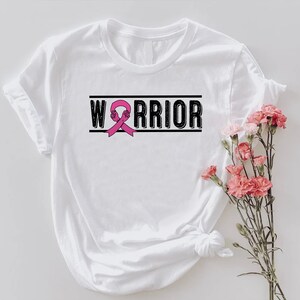 May include: White t-shirt with the word "WARRIOR" in black block letters, with a pink ribbon graphic. The shirt has rolled-up sleeves and is styled with a bouquet of pink flowers.