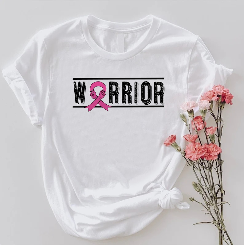 May include: White t-shirt with the word "WARRIOR" in black block letters, with a pink ribbon graphic. The shirt has rolled-up sleeves and is styled with a bouquet of pink flowers.