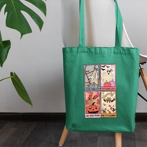 May include: A green tote bag with a Halloween-themed design featuring four illustrated cards. The cards depict a ghost, a pumpkin, a witch, and a friendly ghost. The text on the cards reads "The Ghostly One", "The Ghostly Girl", "The Trick Treat", and "Friendly Ghost".