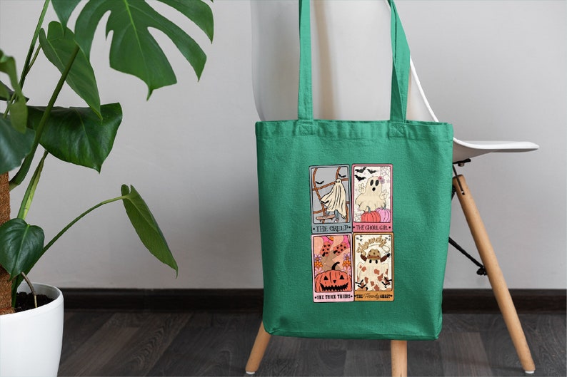 May include: A green tote bag with a Halloween-themed design featuring four illustrated cards. The cards depict a ghost, a pumpkin, a witch, and a friendly ghost. The text on the cards reads "The Ghostly One", "The Ghostly Girl", "The Trick Treat", and "Friendly Ghost".
