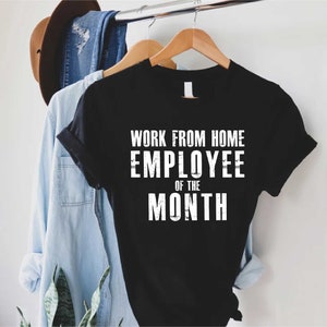 May include: Black t-shirt with the text "WORK FROM HOME EMPLOYEE OF THE MONTH" in white block letters. The shirt is hanging on a wooden hanger with other clothing items in the background.