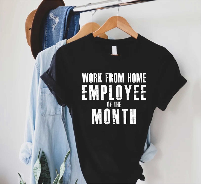 May include: Black t-shirt with the text "WORK FROM HOME EMPLOYEE OF THE MONTH" in white block letters. The shirt is hanging on a wooden hanger with other clothing items in the background.