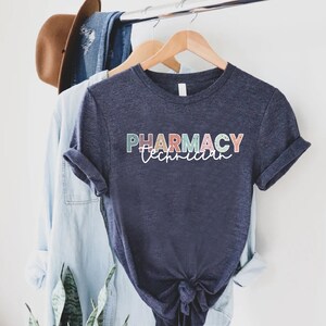 May include: A navy blue t-shirt with the text "PHARMACY Technician" in colorful letters. The letters are outlined in white and have a speckled texture.