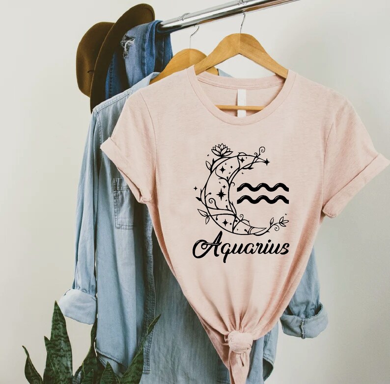 May include: A pink t-shirt with a black graphic of a crescent moon with a floral design and the word "Aquarius" underneath.