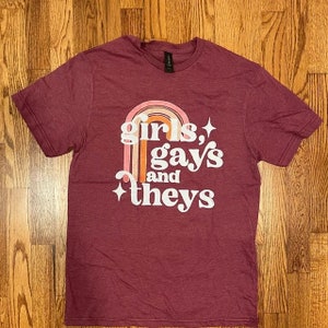 May include: A maroon t-shirt with a rainbow graphic and the text "girls, gays and they's" in white.