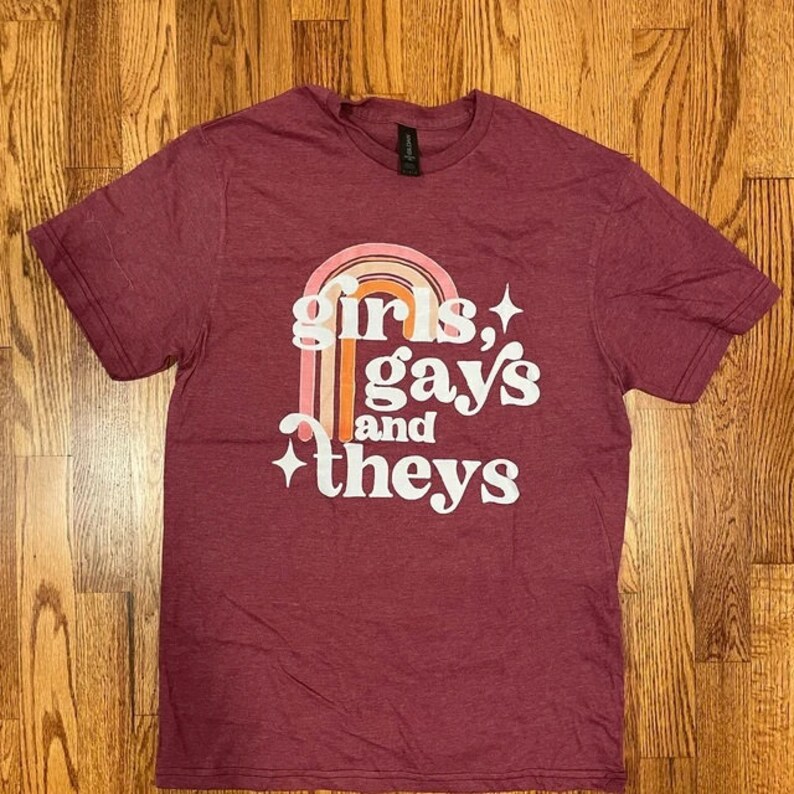 May include: A maroon t-shirt with a rainbow graphic and the text "girls, gays and they's" in white.