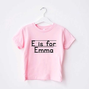 May include: Pink short-sleeved t-shirt with black text that reads "E is for Emma".